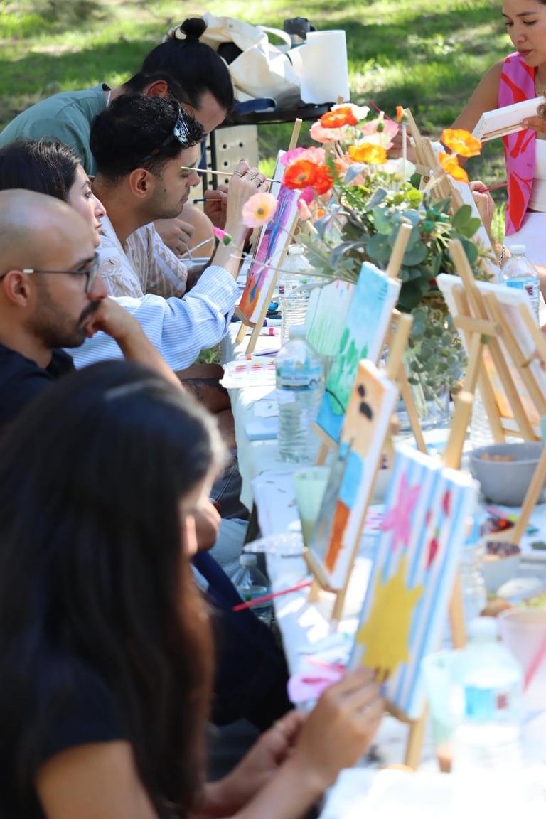 Group of people painting outdoors at a long table with flowers and art supplies on a sunny day