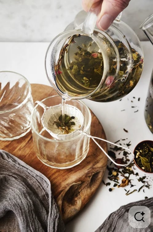 Hand pouring herbal tea from glass teapot into cup with infuser on wooden surface
