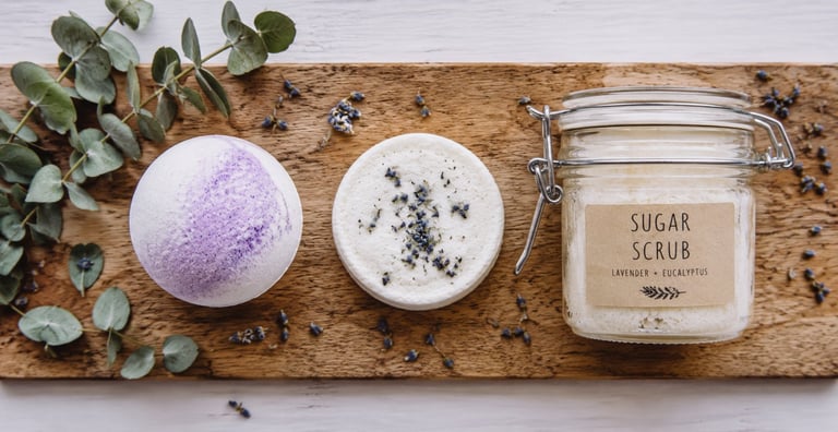 Overhead view of spa products on cork board: purple bath bomb, white scrub with herbs, sugar scrub jar, and eucalyptus leaves